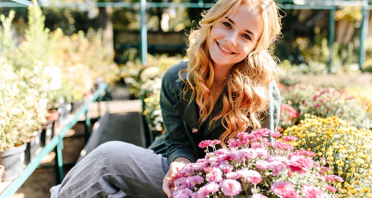 Against background of bright flowering plants and green bushes that grow in garden beautiful blonde girl with green eyes smiles sweetly and basks in sun